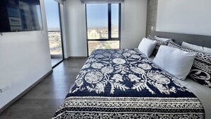 Ein modernes Schlafzimmer mit einem großen Bett, einem an der Wand montierten Fernseher und Blick auf die Stadt durch das Fenster.