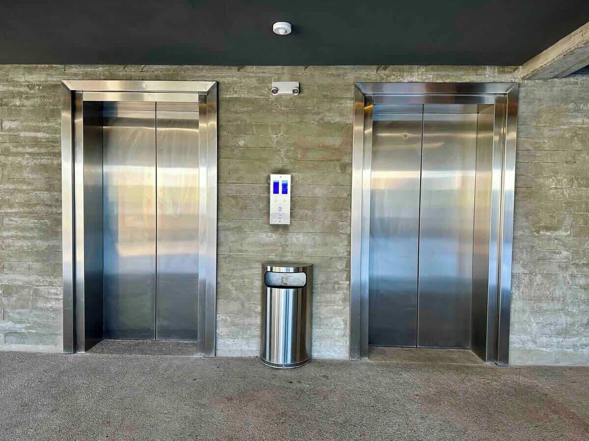 Deux ascenseurs, une poubelle et un panneau de commande fixé au mur, le tout situé dans un hall d’entrée.