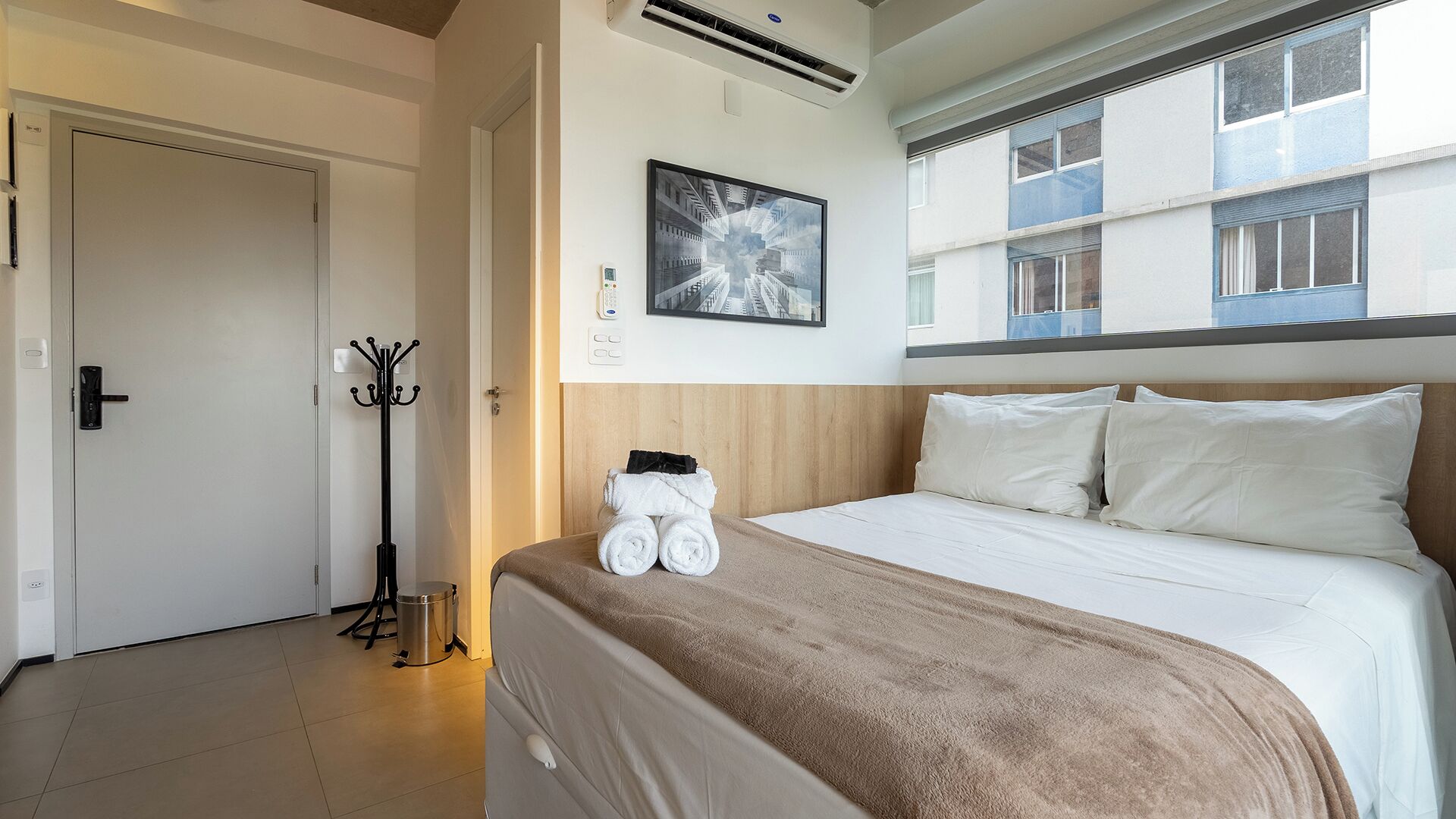 Een hotelkamer met een bed, kapstok, een foto aan de muur en een airconditioning.