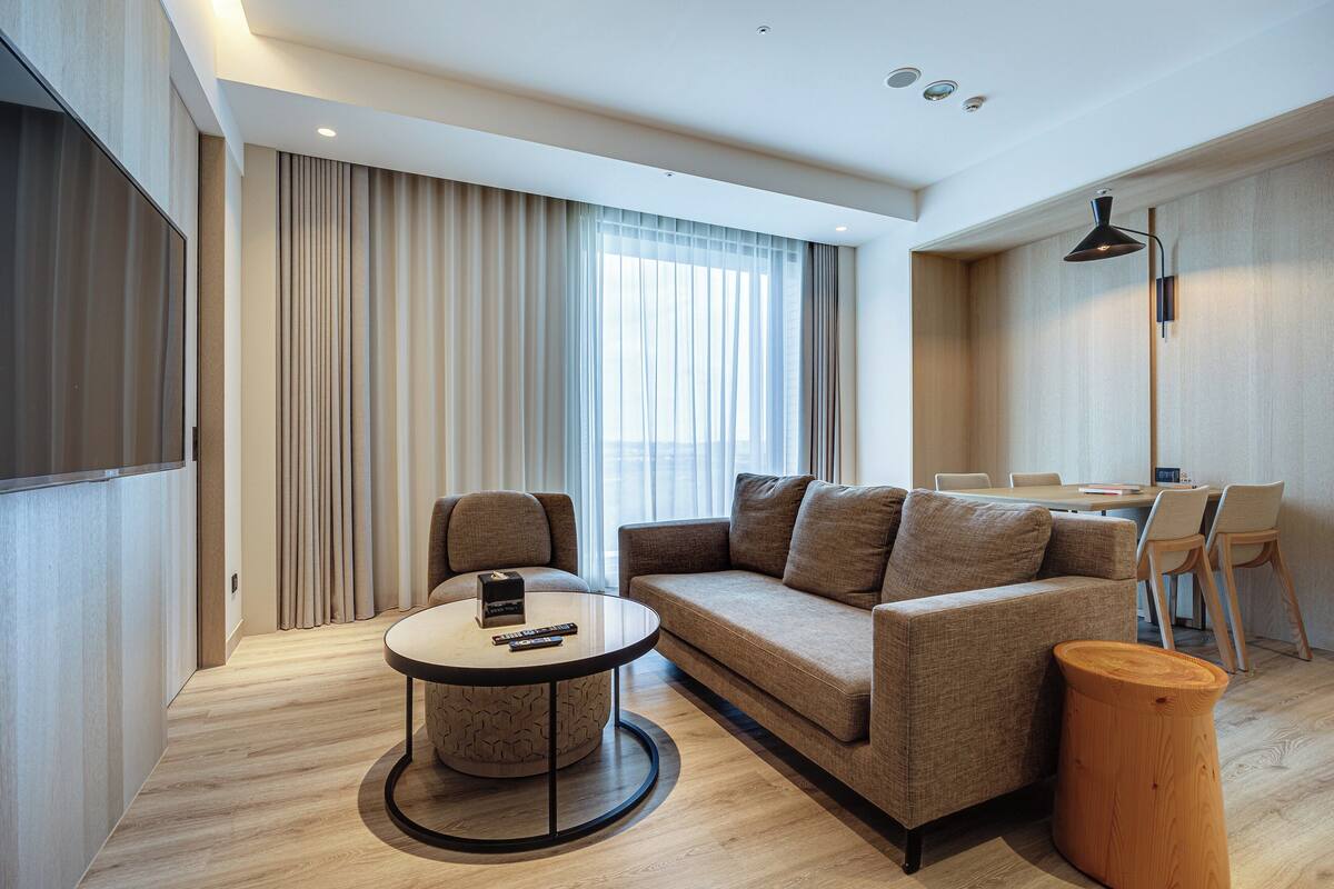 Une chambre d’hôtel moderne avec un canapé, une table basse ronde, une table d’appoint en bois et un coin repas avec des chaises.