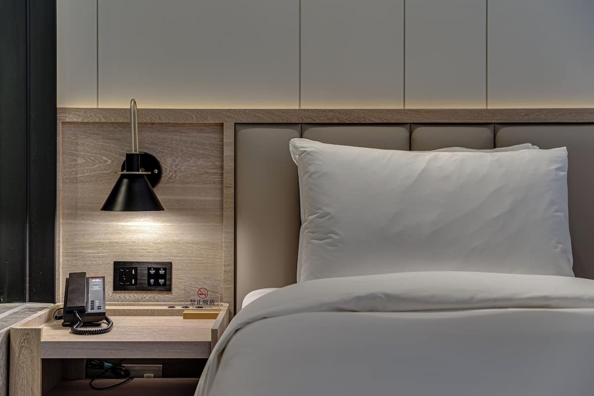 Une chambre d’hôtel comprenant un lit, une table de chevet avec une lampe, un téléphone et un panneau de commande fixé au mur.