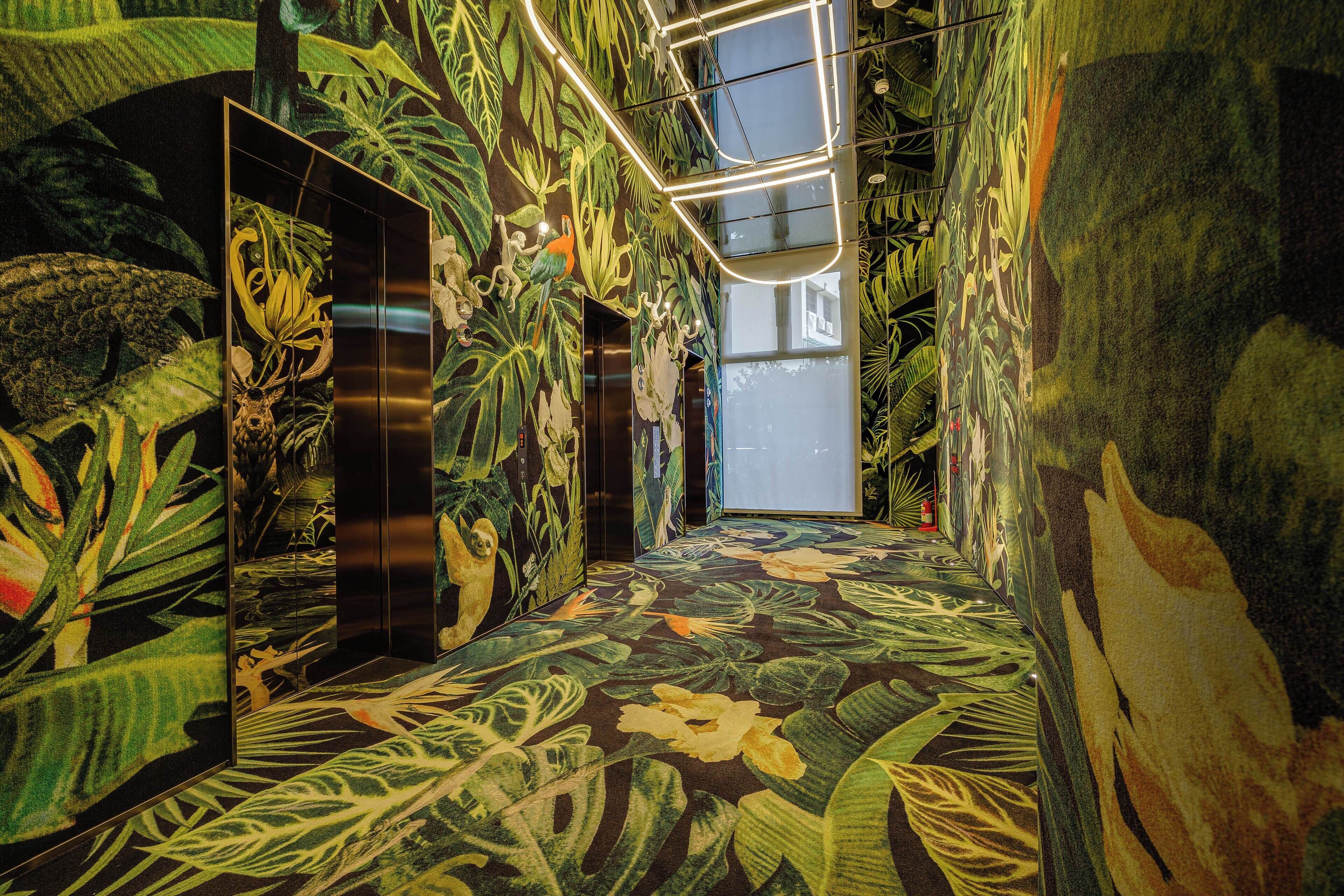 Un pasillo con un mural tropical llamativo, un ascensor de vidrio y vistas al exterior.