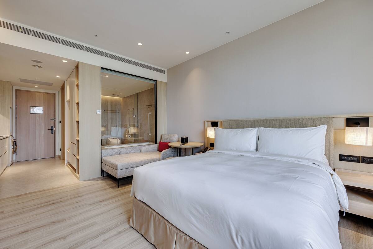Une chambre d’hôtel moderne dotée d’un grand lit, d’un coin salon avec une petite table, d’un dressing et d’une douche en verre.