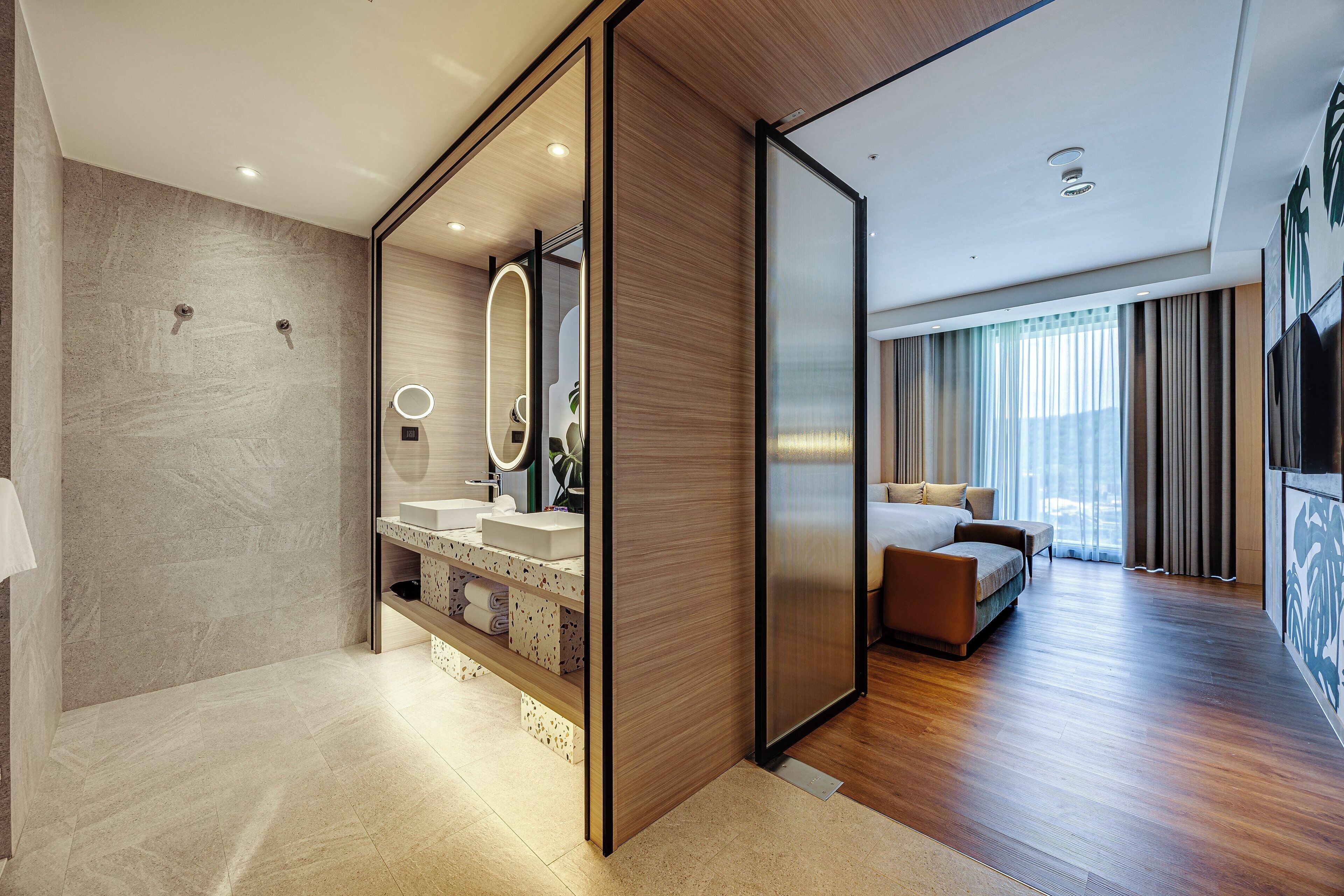 Una habitación de hotel moderna con piso de madera, un espejo grande, lavamanos integrado y una cama con vista a la ciudad.