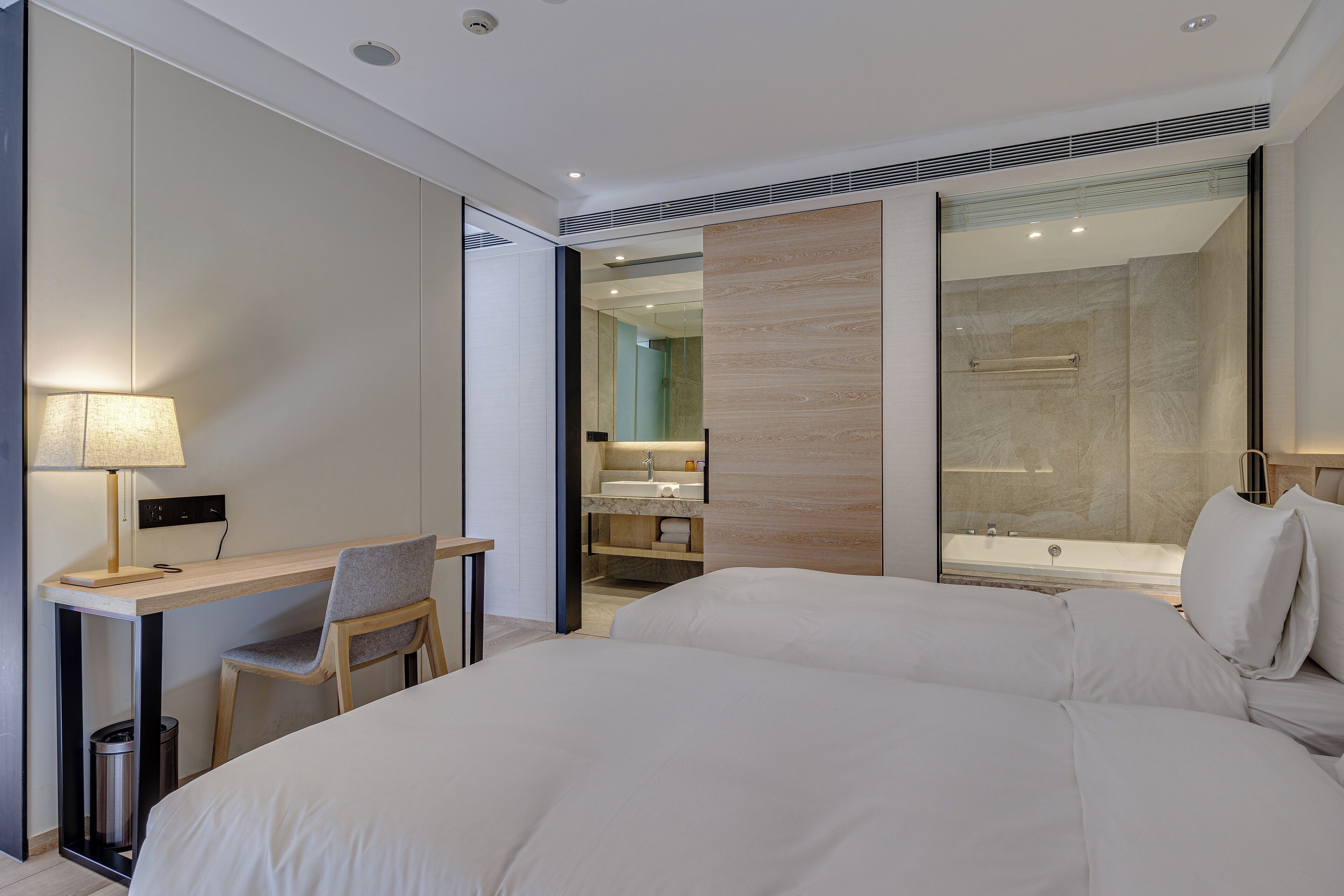 A modern hotel room with a large bed, a desk with a chair, a bathroom with a bathtub, and a large mirror.
