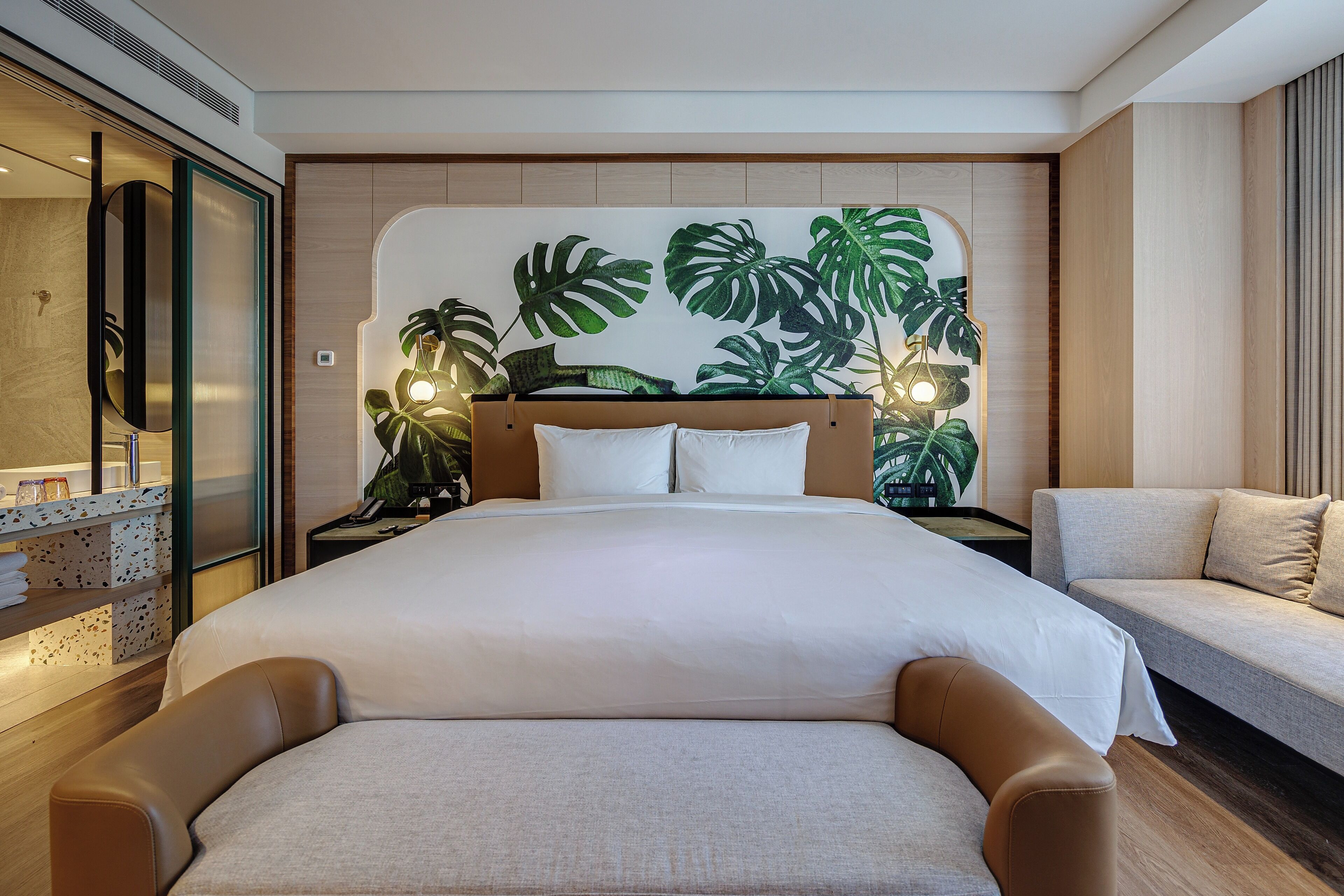 A modern hotel room with a large bed, a sofa, and a wall decorated with tropical leaves.