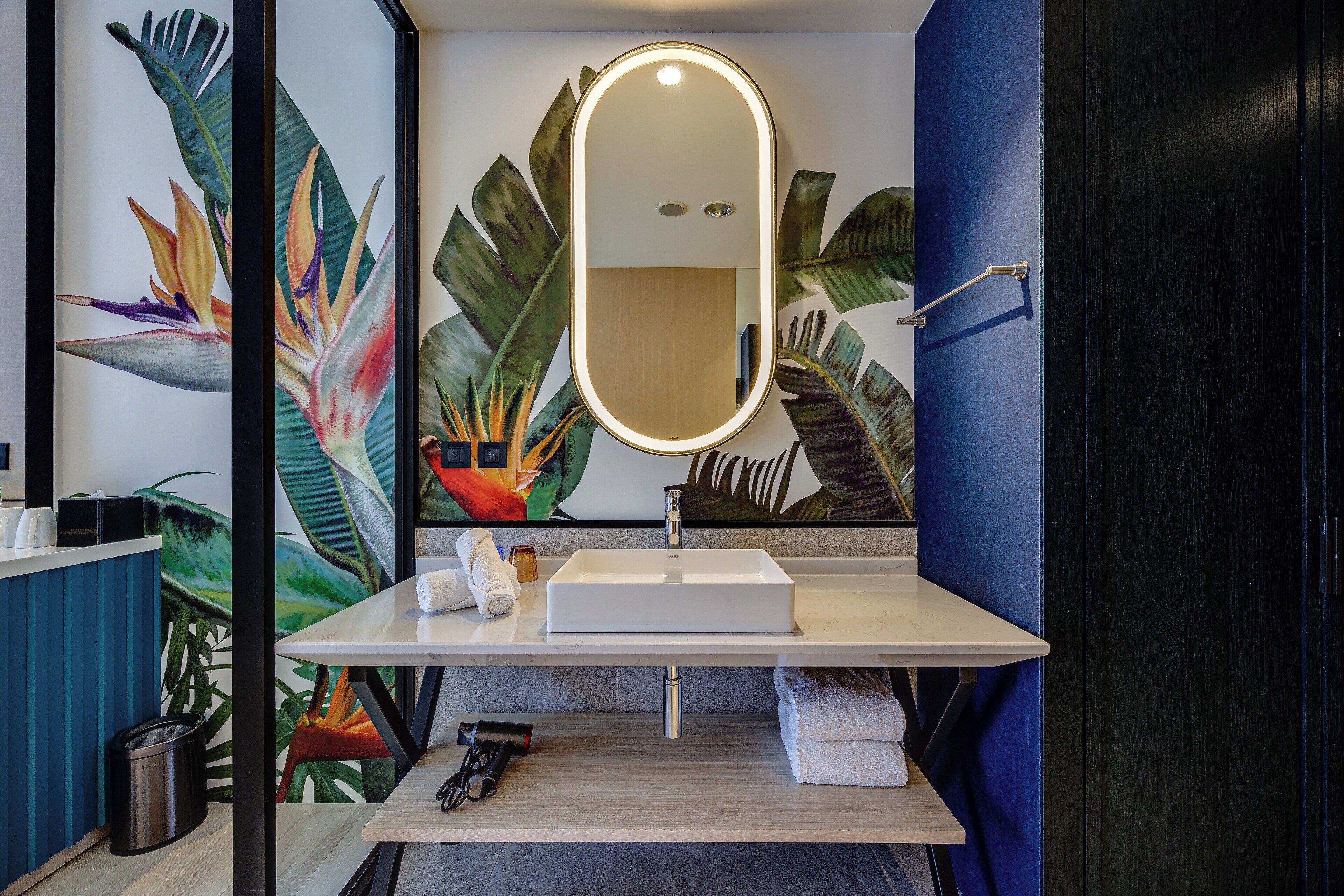 A bathroom with a white sink, a mirror with a light, and a wall with a tropical plant mural.