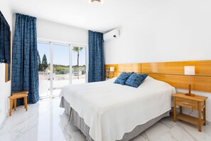 A bedroom with a large bed, wooden headboard, two bedside tables with lamps, a small stool, and a view of the outdoors through a sliding door.