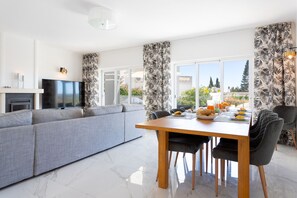 A modern living room with a grey sofa, a dining table with chairs, and a TV on a stand.