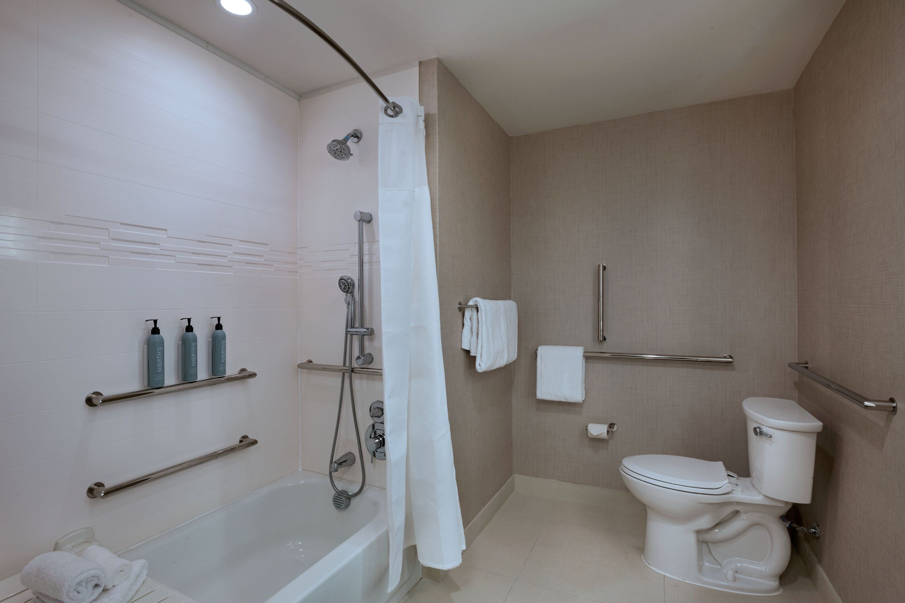 A modern bathroom with a bathtub, toilet, and shower. There are handrails, a towel rack, and toiletries on the wall.