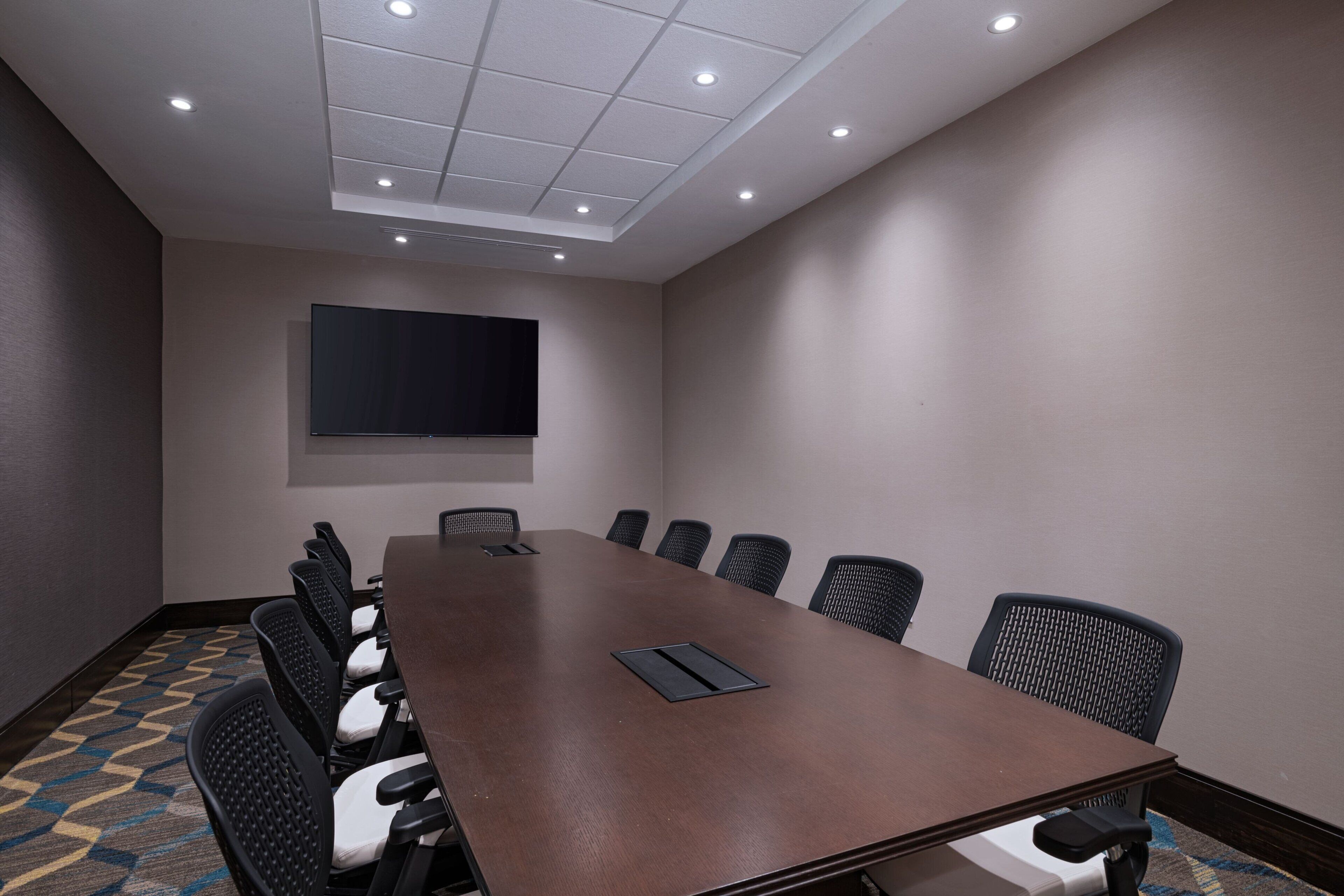 A conference room with a long table, chairs, a flat-screen TV, and recessed ceiling lights.