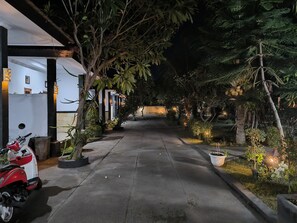 A nighttime view of a pathway lined with potted plants and trees, leading to a building with a white facade and a red scooter parked nearby. - Puri Raka Villa (Seririt)