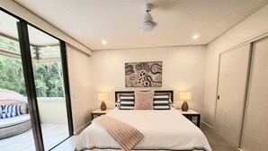 A bedroom with a large bed, two bedside tables with lamps, a ceiling fan, a large window with a view of greenery, and a sliding glass door leading to a balcony. - Luxury Spa Suite #220 Temple Palm Cove (Palm Cove)