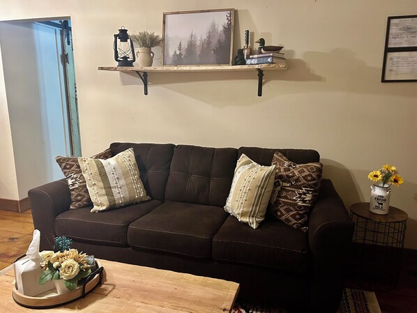 A dark brown leather sofa with patterned cushions, a small wooden table with a tray of flowers, and a shelf above holding a lantern, books, and decorative items. - 🏡Little Bear Cabin In Helen🐻 (Helen)