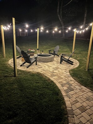 An outdoor patio area with a stone fire pit, two Adirondack chairs, and string lights. - 🏡Little Bear Cabin In Helen🐻 (Helen)