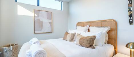 Een keurig opgemaakt bed met witte lakens, een beige plaid, een rieten hoofdbord en een nachtkastje met een lamp.