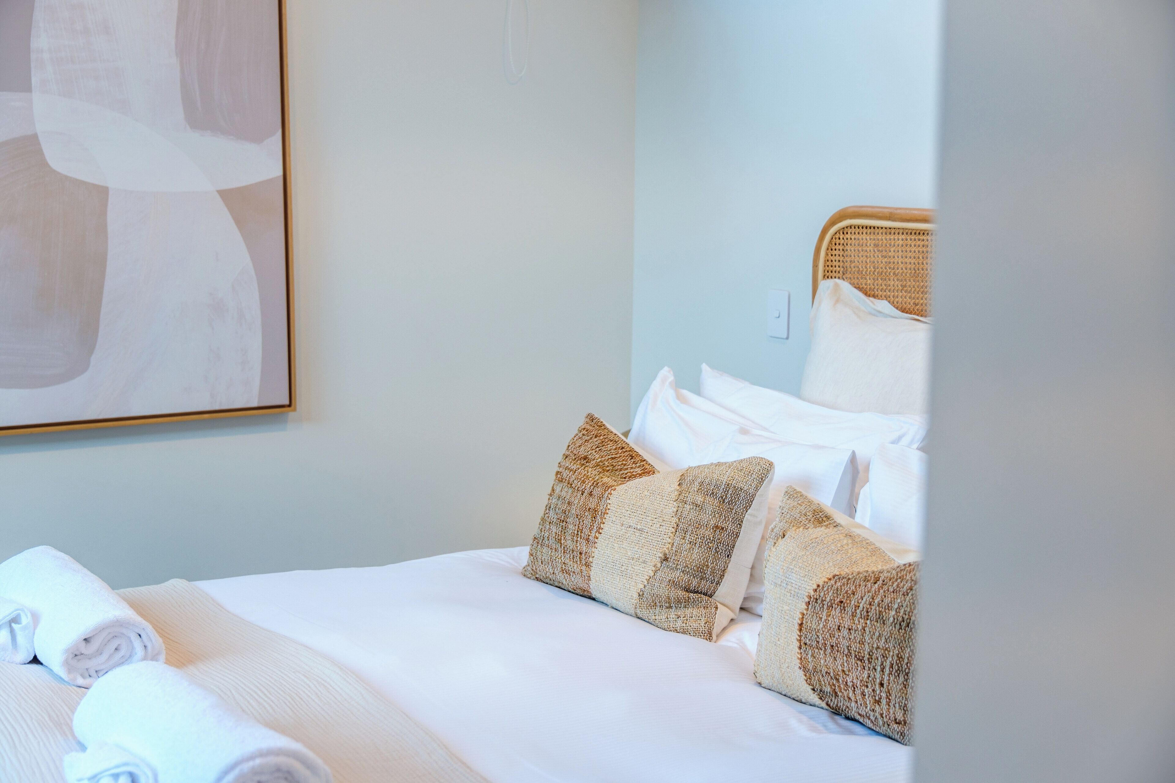 A neatly made bed with white linens and patterned pillows, a wicker headboard, and a large abstract painting on the wall.