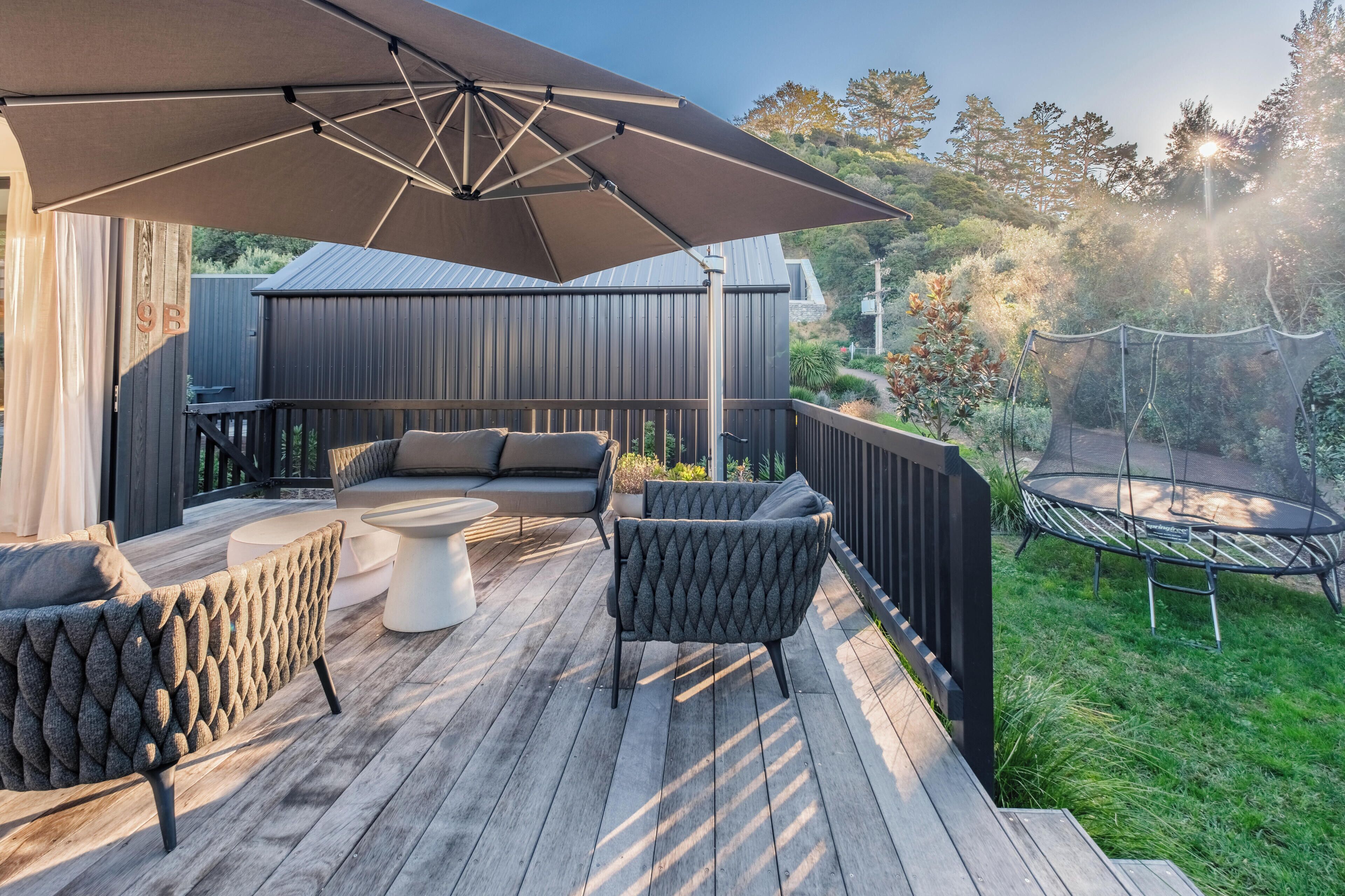Eine Holzterrasse mit Gartenmöbeln, einem großen Sonnenschirm und einem Trampolin.