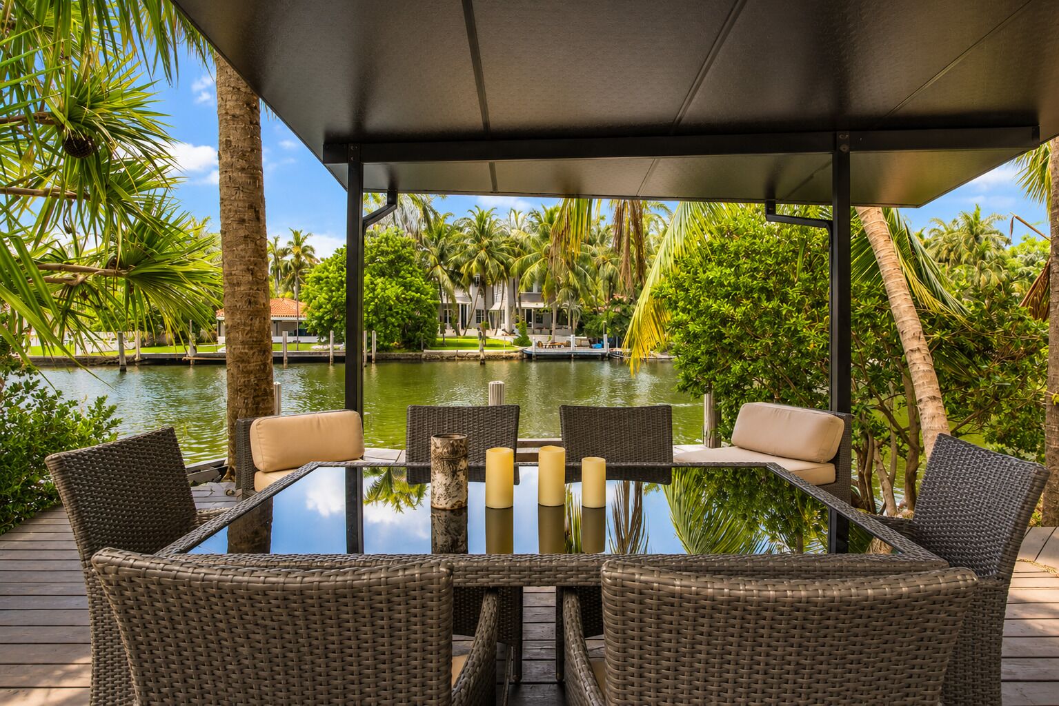 Een glazen tafel met rieten stoelen en kaarsen, met uitzicht op een waterpartij met palmbomen en een steiger.