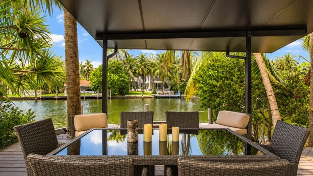 A glass-top table with wicker chairs and candles, overlooking a body of water with palm trees and a dock.
