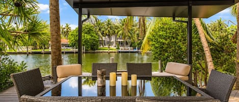 Una mesa de vidrio con sillas de mimbre y velas, con vista a una masa de agua con palmeras y un muelle.