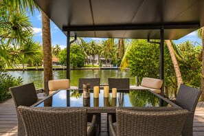Una mesa de vidrio con sillas de mimbre y velas, con vista a una masa de agua con palmeras y un muelle.