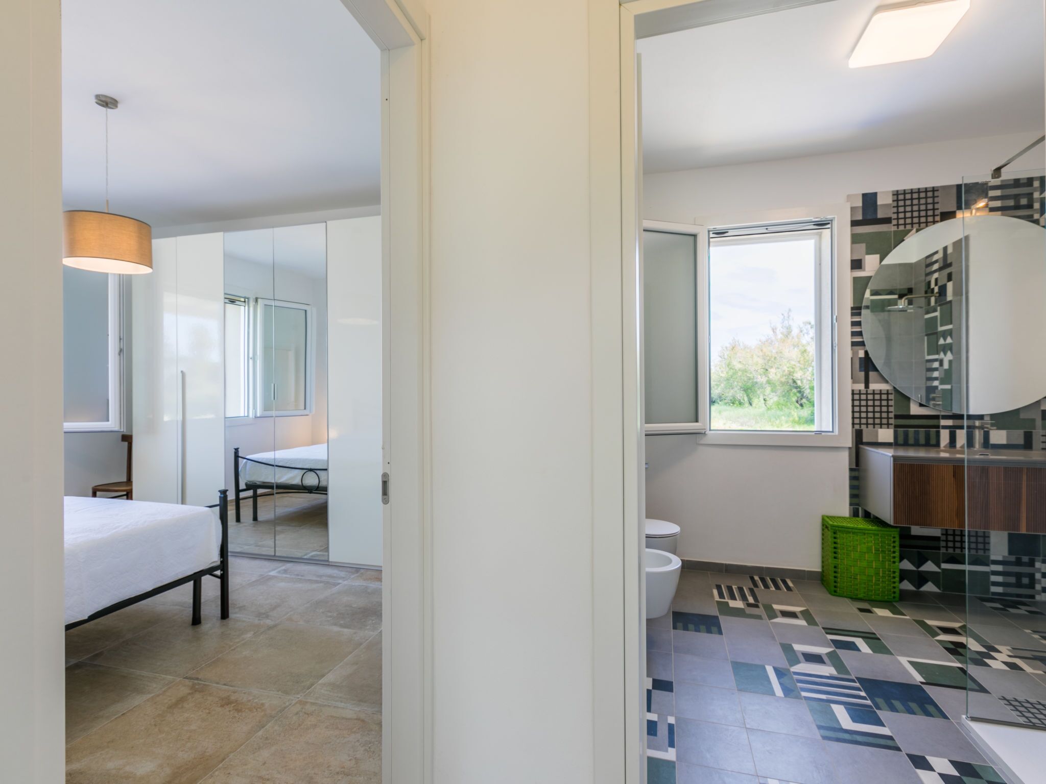 A modern bathroom with a geometric tile floor, a large mirror, and a window providing natural light.