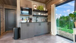 A modern kitchen with grey cabinets, a black microwave, and a large window offering a view of greenery. - Hart's Overlook | Hot Tub, Stunning Mtn Views + Private Pond! (Augusta)