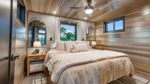A bedroom with a wooden ceiling, a bed with a white comforter, a nightstand with a lamp, and a window with a view of greenery. - Hart's Overlook | Hot Tub, Stunning Mtn Views + Private Pond! (Augusta)