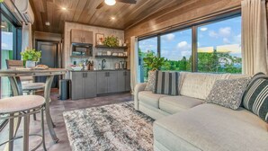 A modern kitchen with wooden cabinetry, a dining area with bar stools, and a large window offering a view of greenery. - Hart's Overlook | Hot Tub, Stunning Mtn Views + Private Pond! (Augusta)