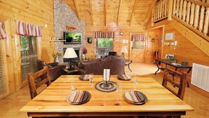 Interior acogedor de una cabaña con una mesa de comedor para cuatro, una chimenea de piedra y un dormitorio en un entrepiso con escalera de madera.