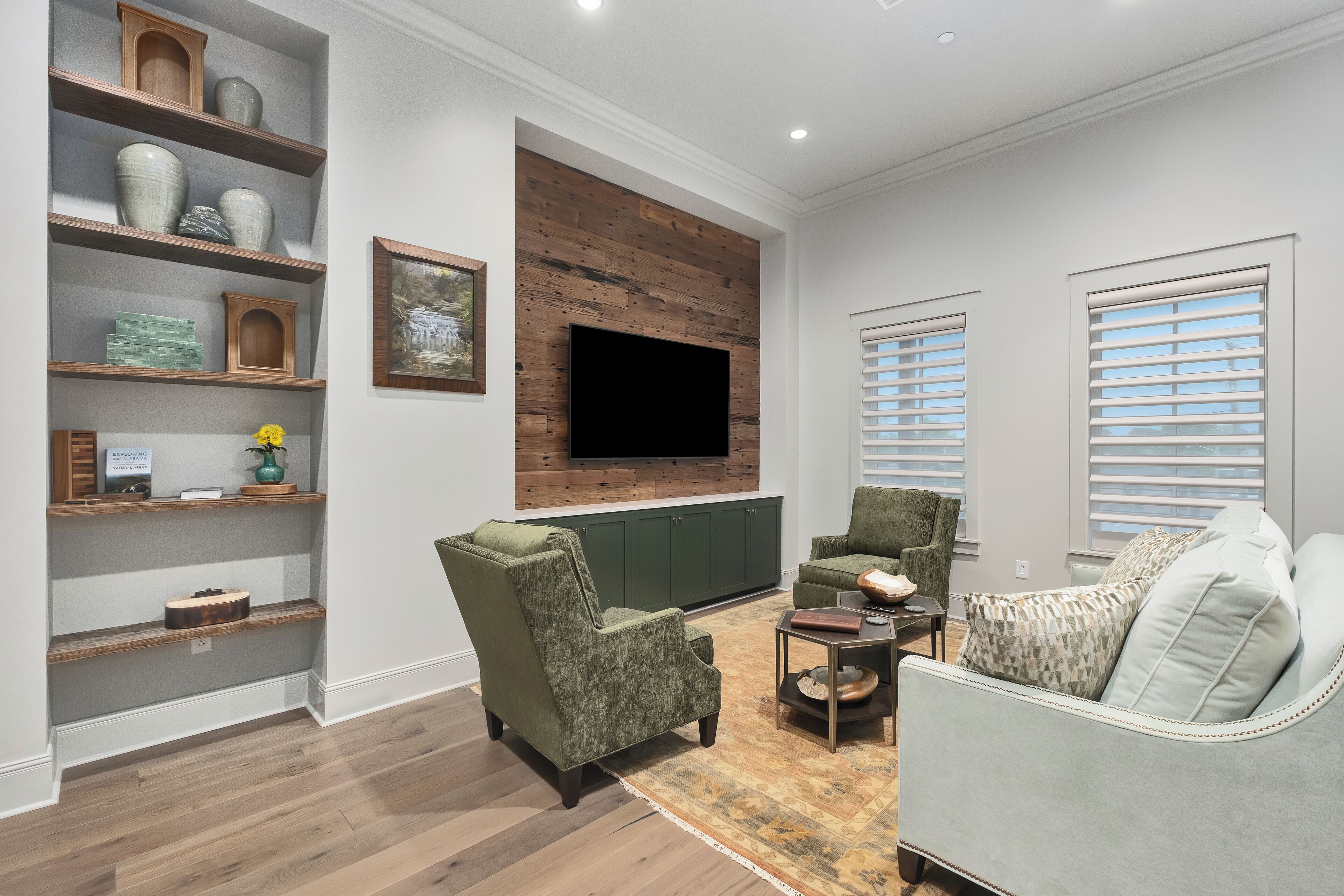 Una sala de estar con una unidad mural de madera, un televisor de pantalla plana, un sillón verde, un sofá gris claro y una alfombra estampada.