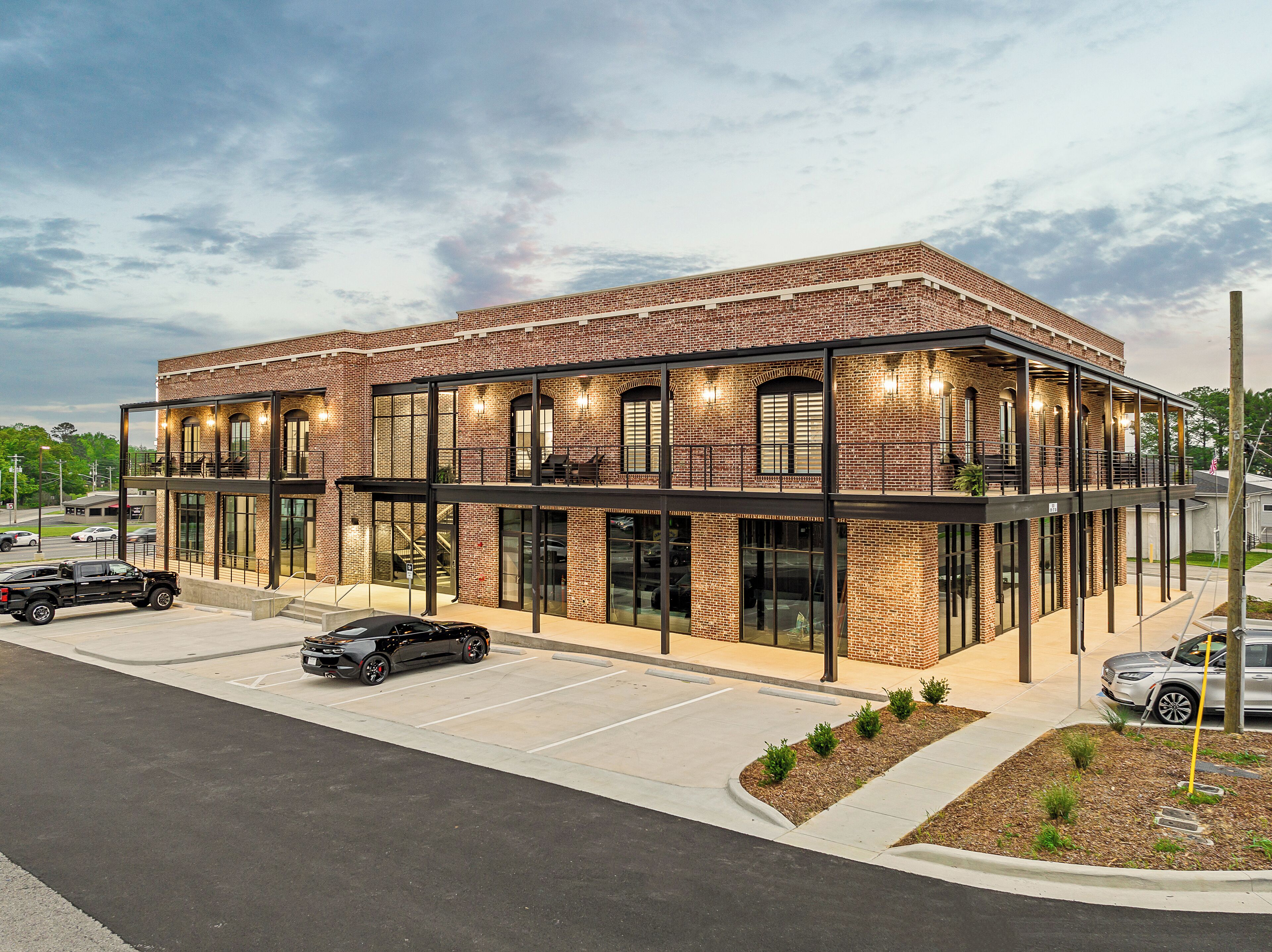 Un bâtiment moderne à la façade en briques et aux grandes fenêtres, doté d’un espace extérieur couvert avec des sièges et d’un parking.