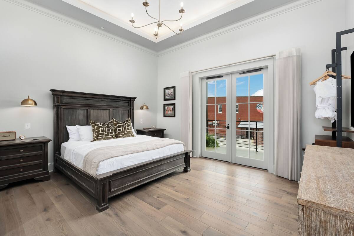 A bedroom with a large bed, dark wooden headboard, two bedside tables with lamps, a sliding glass door to a balcony, and a wooden dresser.