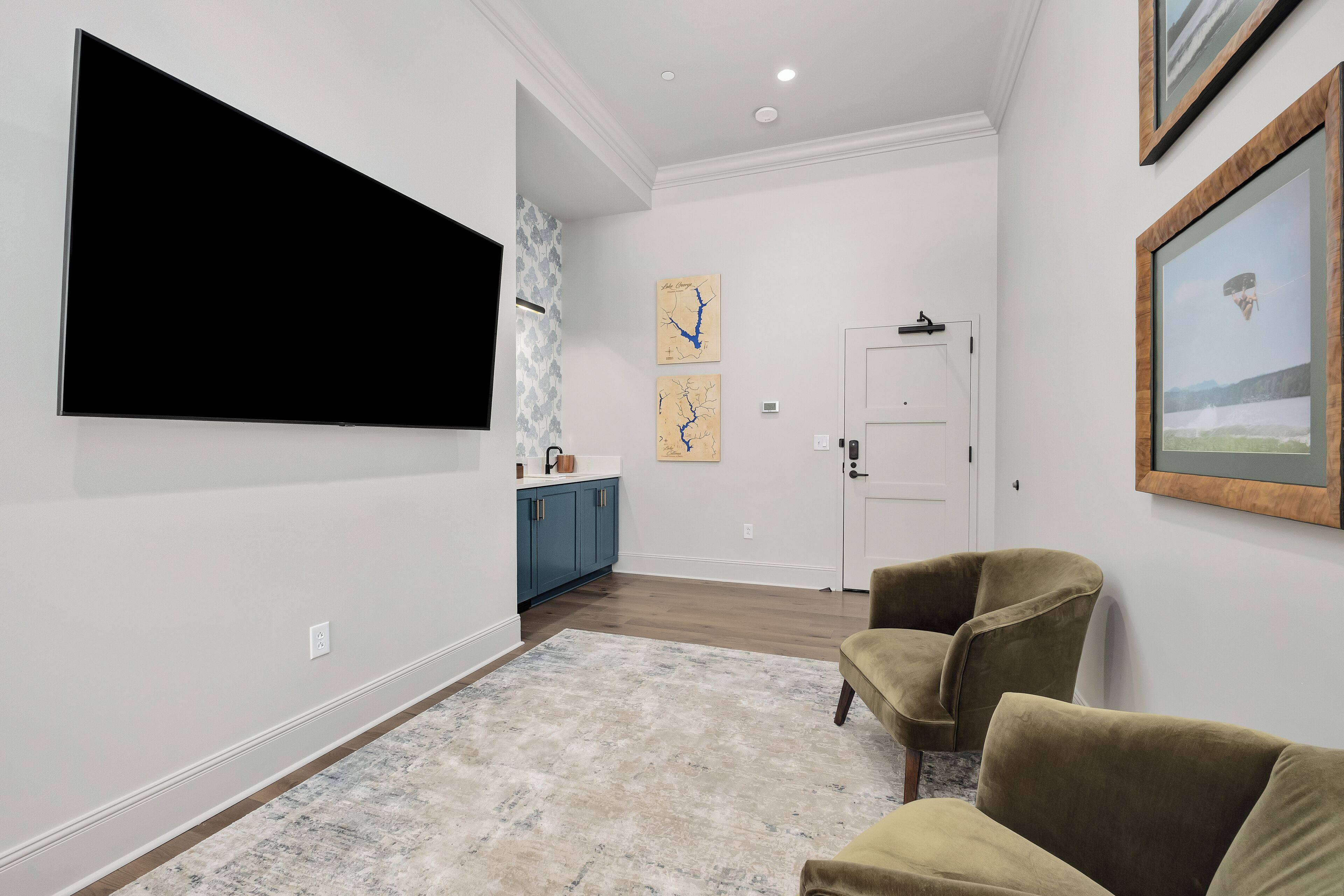 Een moderne woonkamer met een flatscreen televisie aan de muur, een gezellige zithoek met twee fauteuils en een keuken met blauwe kastjes en een wit aanrecht.