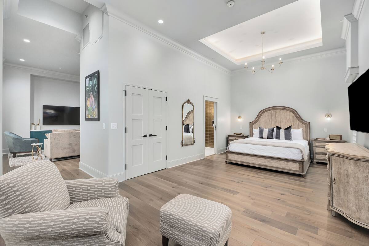 A spacious bedroom with a large bed, a sitting area with a chair and ottoman, a TV, and a mirror.