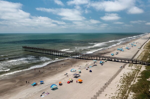 A beach with a pier extending into the ocean, umbrellas, and beachgoers.