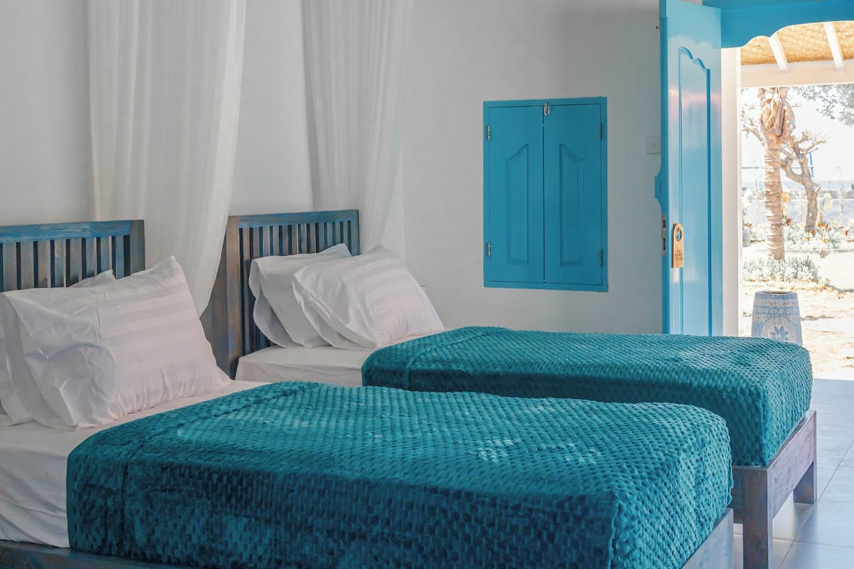 A bedroom with two beds, white curtains, a blue door, and a view of the outdoors.