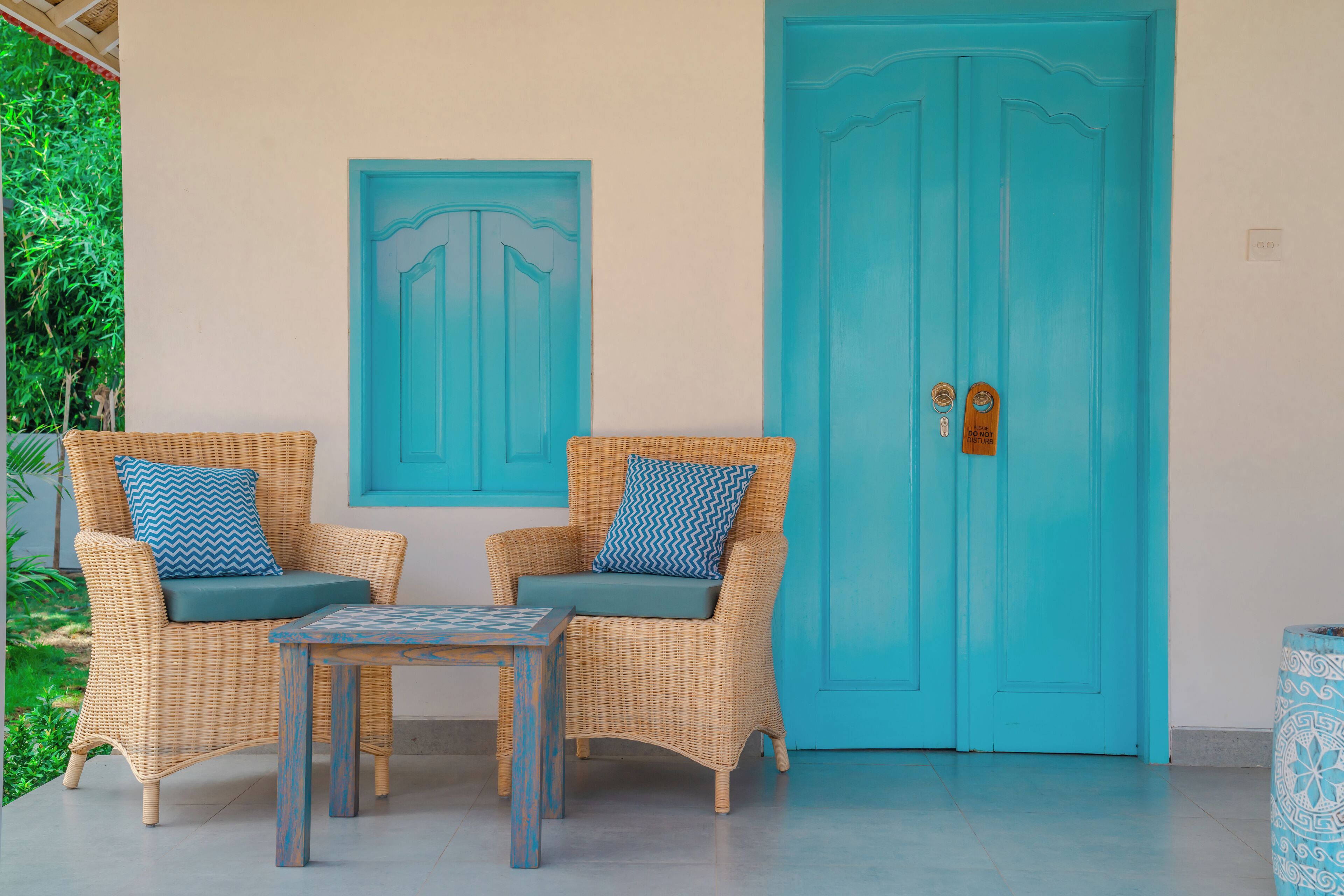 Two wicker chairs with blue cushions and a small table, a turquoise door, and a decorative vase.