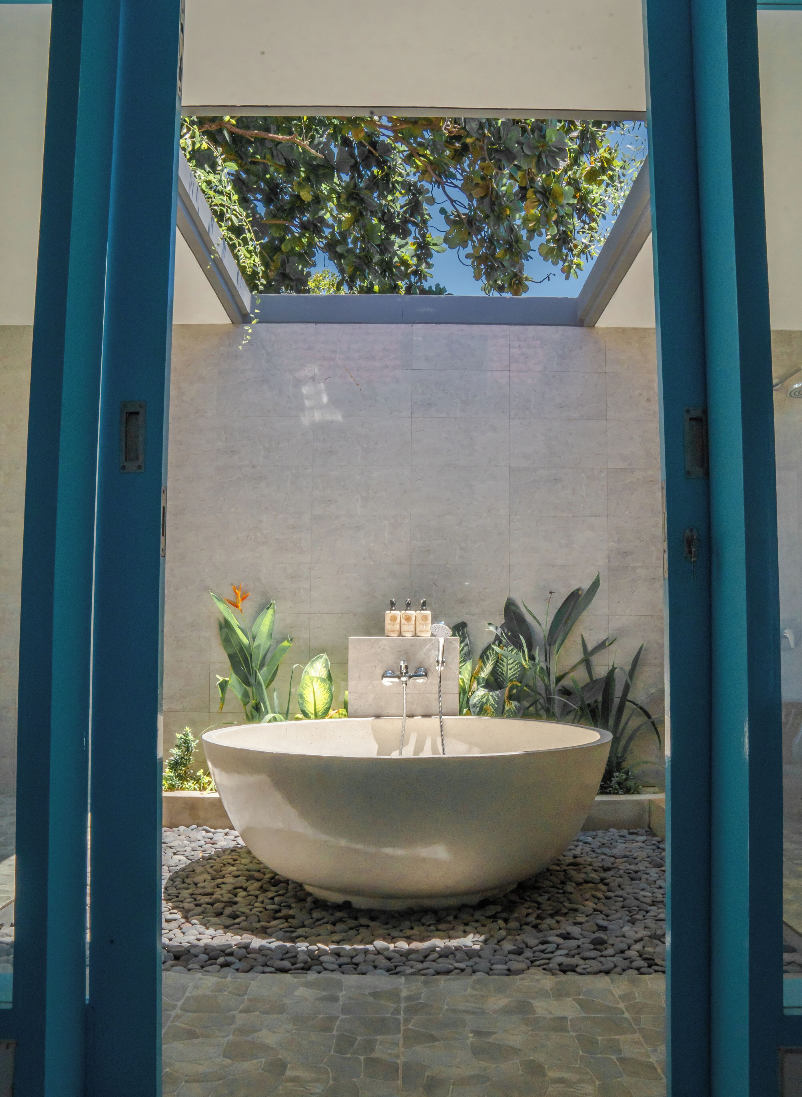 A large outdoor bathtub with a stone surround, surrounded by plants and a tiled wall.