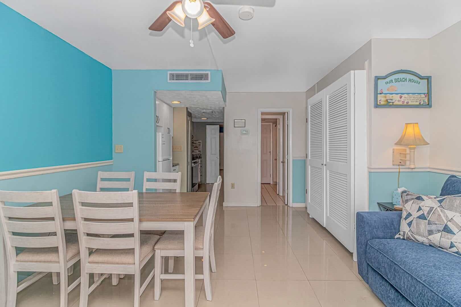 A room with a dining table, chairs, a ceiling fan, a sofa, and a wall sign.