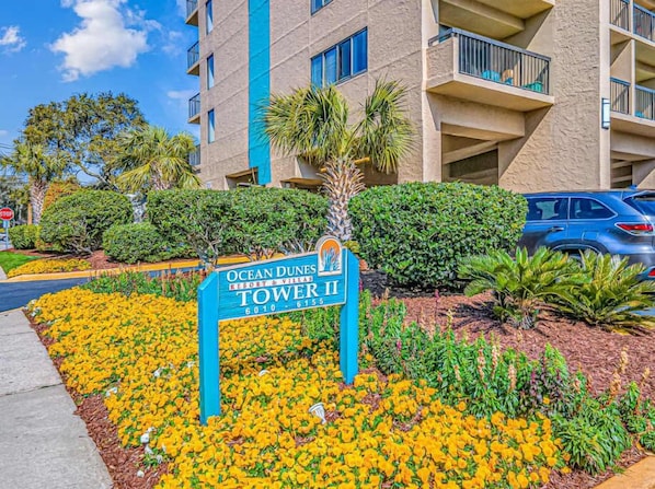 A sign for Ocean Dunes Tower II, a multi-story building with balconies, surrounded by landscaped gardens and parked cars.