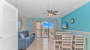 Une pièce avec un mur bleu, un ventilateur de plafond, une table à manger avec des chaises, un canapé avec des coussins et une vue sur l’extérieur, visible à travers une porte vitrée.