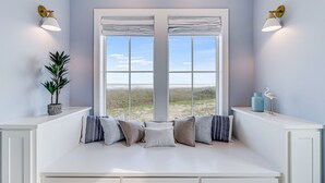 A room with a large window, a white bench with cushions, a potted plant, and wall-mounted lights. - Newly built oceanfront home with elevator, balconies, and awe-inspiring views (Port Aransas)