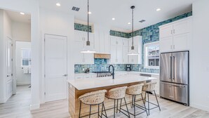 A modern kitchen with white cabinets, a stainless steel refrigerator, and a bar area with wicker stools. - Newly built oceanfront home with elevator, balconies, and awe-inspiring views (Port Aransas)