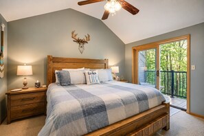 Une chambre avec un grand lit, une tête de lit en bois, deux tables de chevet avec des lampes, une décoration murale représentant une tête de cerf et un balcon avec vue sur les arbres.