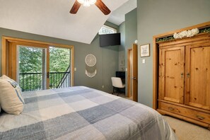 Une chambre avec un grand lit, une armoire en bois, un bureau avec une chaise, un téléviseur fixé au mur et un balcon avec vue sur les arbres.