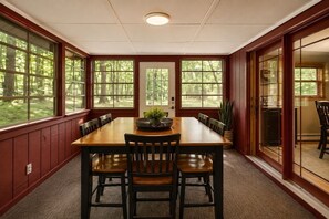 Comedor con mesa y sillas de madera, ventanas amplias con vista a árboles y una puerta que da a otra habitación.