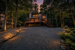 A two-story wooden cabin with a deck, surrounded by a gravel driveway and trees. - New Listing: On the Rocks in the Hideout (Lake Ariel)