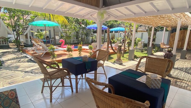A covered outdoor seating area with wicker chairs and tables, a pool in the background, and a tropical garden setting.